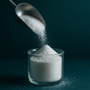 A creative close-up of fine white phosphate powder being poured from a stainless steel scoop into a clear glass beaker, with granules captured mid-air against a dark teal background and soft studio lighting.